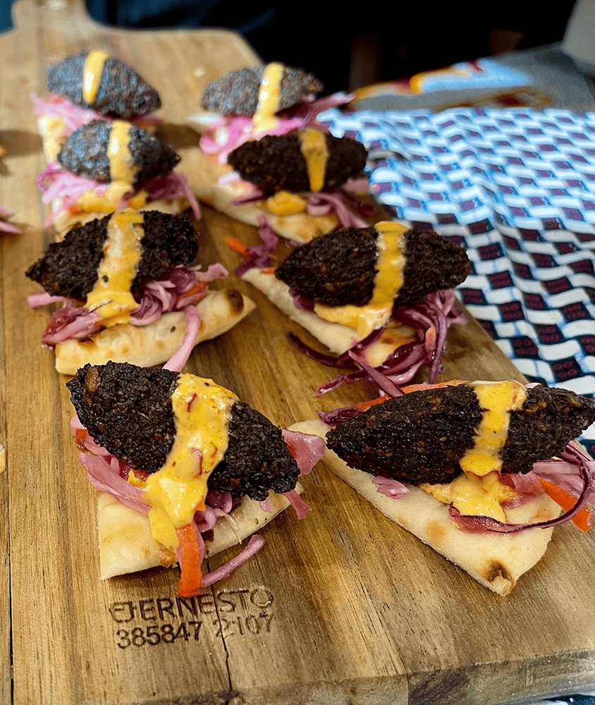 A wooden board with small pieces of flatbread topped with vegetables and a dark crispy topping, drizzled with yellow sauce.