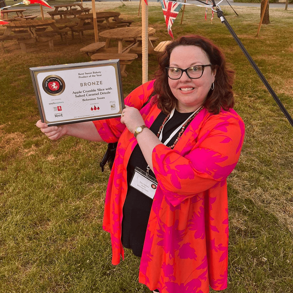 A woman smiling and holding a framed certificate while standing outdoors, with picnic tables and Union Jack bunting visible in the background.