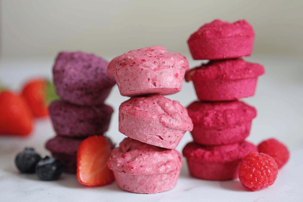 Stacks of colourful fermented macarons from Fermenti in shades of purple, pink, and red, surrounded by fresh fruits including strawberries, raspberries, and blueberries.